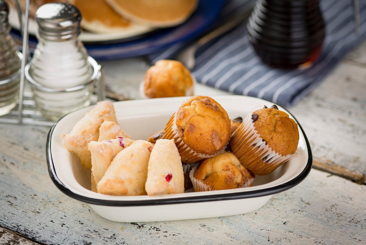 A white Enamelware Serving Pan holds blueberry muffins in paper liners and sugar-dusted scones. In the background are salt and pepper shakers, pancakes on a blue plate, and a syrup bottle atop a striped cloth. Dishwasher safe for easy cleanup.