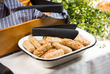 A white enamelware serving pan filled with cooked sausage links sits on a tablecloth beside black tongs. In the background, a wooden basket lined with a blue-striped cloth and green leafy decor completes the scene.