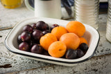 A white porcelain enamel coated steel Enamelware serving pan filled with fresh dark purple plums and bright orange apricots sits on a rustic white wooden table. Blurred mugs and jars are visible in the background.