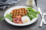 A porcelain enamel plate holds two brown waffles, a poached egg topped with black pepper, prosciutto slices, and fresh arugula. A fork, knife, napkin, and small pitcher of cream rest nearby on the gray tabletop.