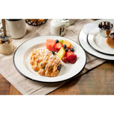 A white porcelain enamel plate holds a fruit Danish pastry drizzled with icing and a colorful serving of cut watermelon, pineapple, cantaloupe, strawberries, and blueberries. The plate sits on a beige cloth with yogurt and other breakfast items nearby.