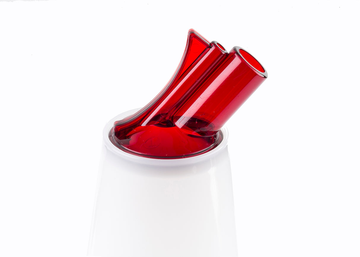 A close-up of a modern, abstract red and clear glass object with two tilted, color-coded pour spouts, set atop a smooth white base against a plain white background. The sleek, minimalist design suggests a contemporary decorative or functional BPA free item.