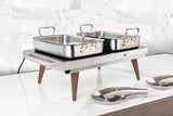 A stainless steel buffet roaster with wooden legs holds two 6 qt chafing dishes. The rectangular warming tray sits on a white marble countertop before a light gray tiled wall, with serving spoons resting on a nearby oval tray.