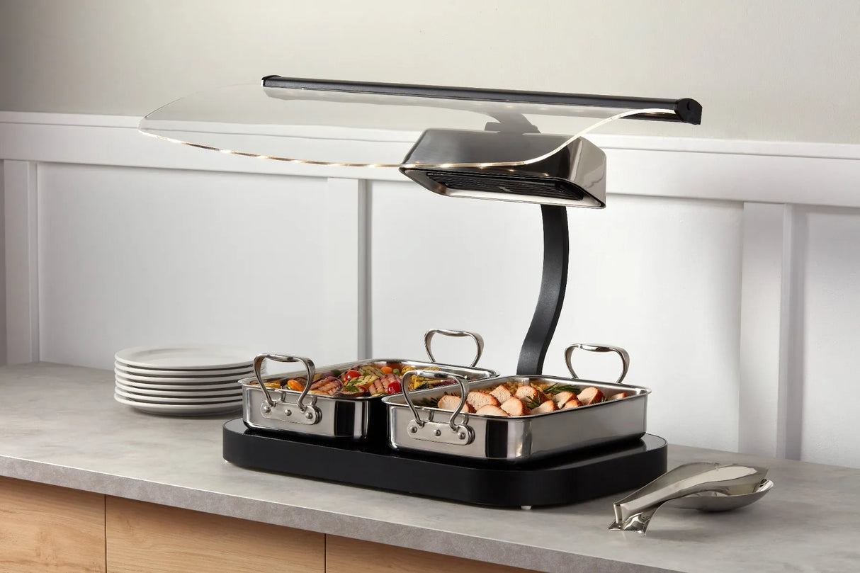 A buffet-style countertop Buffet Roaster with two 3 qt stainless steel trays filled with assorted cooked food sits under a heat lamp. Plates are stacked to the left, tongs rest on the right, all on a light gray counter against a white paneled wall.