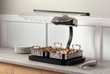 A buffet-style countertop Buffet Roaster with two 3 qt stainless steel trays filled with assorted cooked food sits under a heat lamp. Plates are stacked to the left, tongs rest on the right, all on a light gray counter against a white paneled wall.