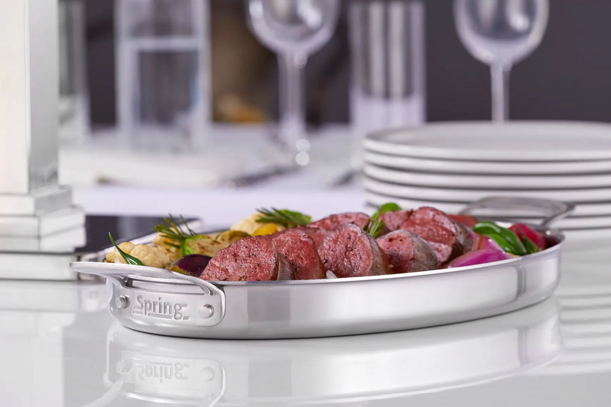 A 1-1/2 qt. oval buffet sauté stainless steel serving dish filled with sliced cooked meat, grilled vegetables, and fresh herbs sits on a reflective white table. Stacks of plates and wine glasses blur in the elegant dining background.