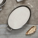 An empty, oval stainless steel serving tray with handles rests on a burlap cloth, perfect for buffet sauté presentations. Nearby, a metallic gold, leaf-shaped dish sits on a rustic wooden surface, with another metal dish peeking from the top.