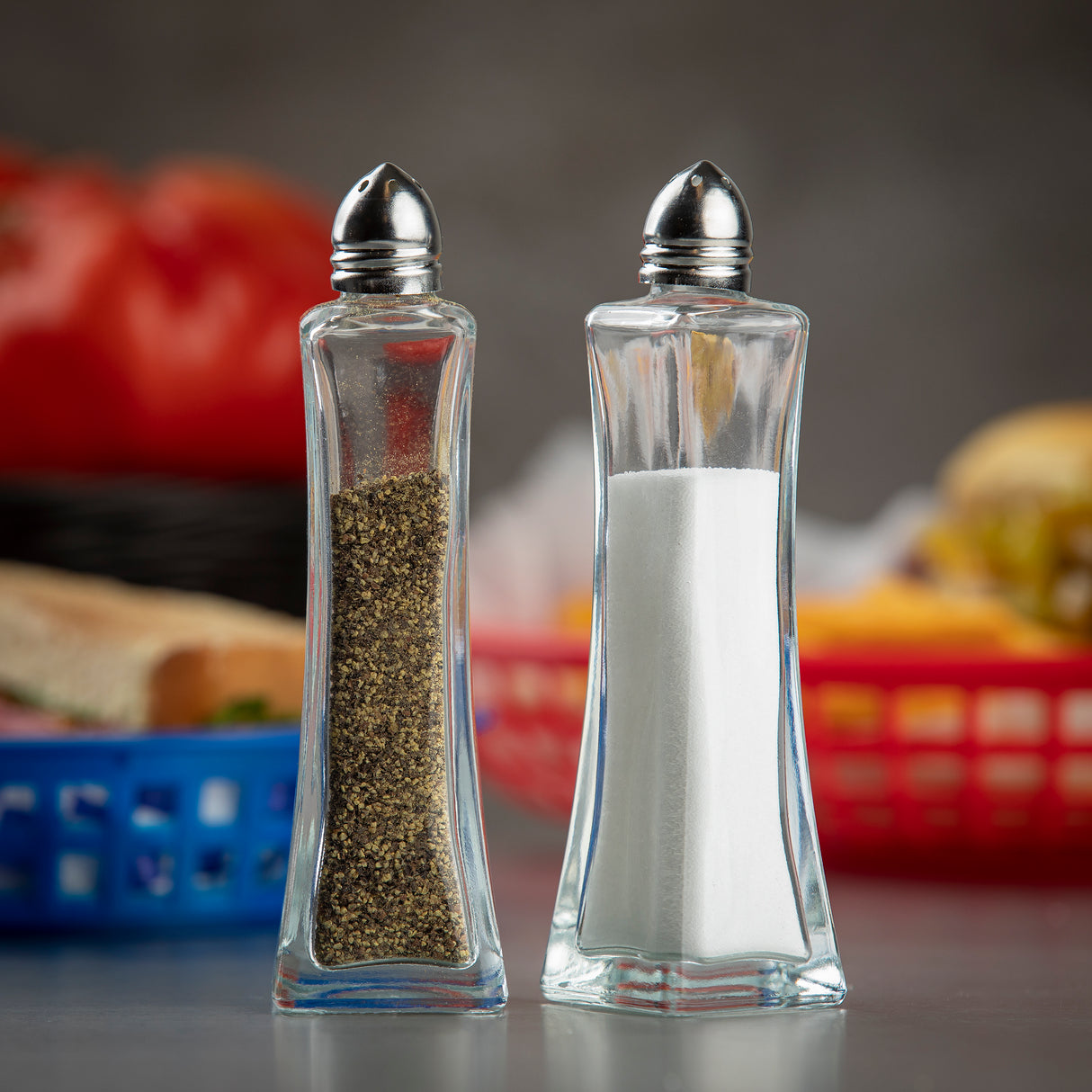 Two Salt & Pepper Shakers with a glass body and silver tops stand side by side on a tabletop. Black pepper fills the left shaker, white salt the right. Blurred food items, including a sandwich and tomatoes, are in the background. Dishwasher safe.