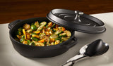 A black Ironlite Casserole filled with roasted vegetables—sliced zucchini, yellow squash, and Brussels sprouts—sits on a white countertop. Perfect for induction cooking, its lid is tilted open with a serving spoon and tongs beside it.