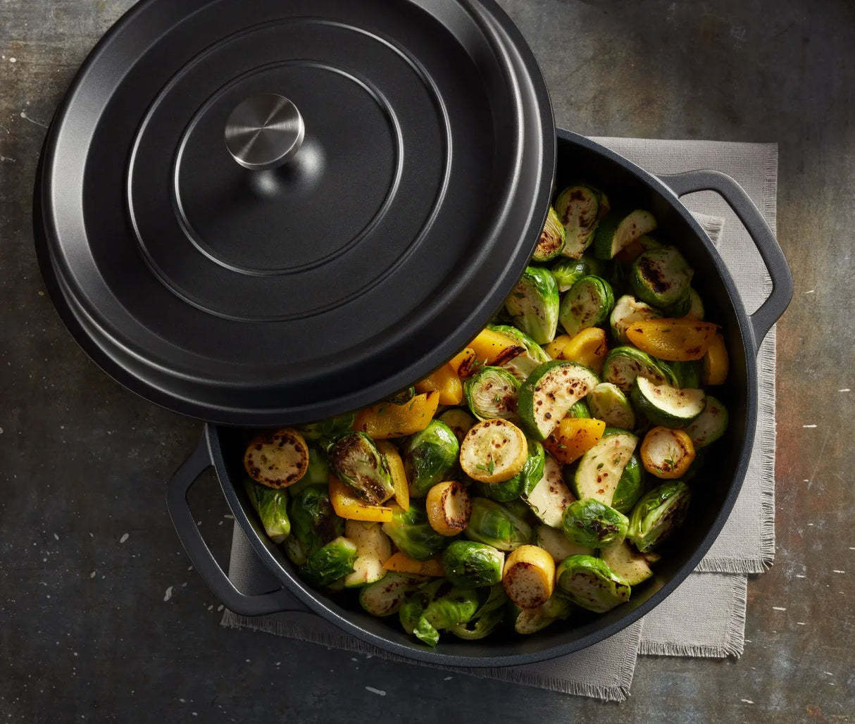 A black Ironlite Casserole pot, partially covered with a lid, sits on a gray cloth. Inside are roasted Brussels sprouts and yellow squash, lightly browned and seasoned with herbs—the pot ready for induction cooking on a dark, rustic surface.