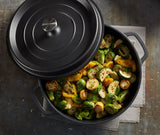 A black Ironlite Casserole pot, partially covered with a lid, sits on a gray cloth. Inside are roasted Brussels sprouts and yellow squash, lightly browned and seasoned with herbs—the pot ready for induction cooking on a dark, rustic surface.