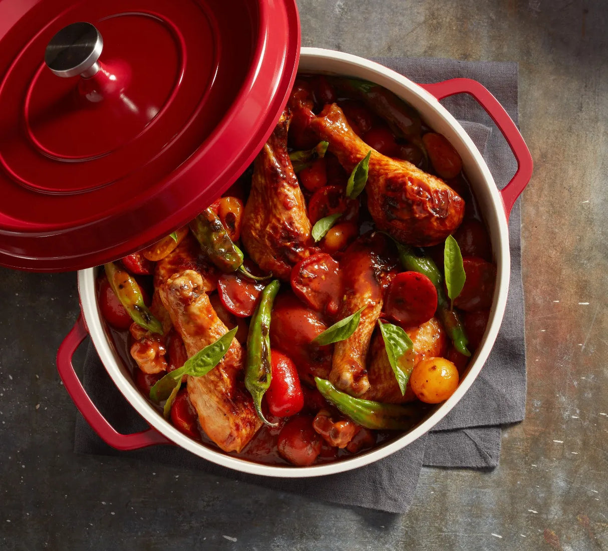 A red Ironlite Casserole filled with cooked chicken drumsticks, cherry tomatoes, green beans, and fresh basil sits on a dark cloth napkin atop a rustic table. The lid is partially open, showing the colorful, saucy contents inside. Perfect for induction cooking.