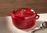 A shiny red Ironlite Casserole Dutch oven with a lid and metal knob sits on a glossy white countertop. The Dutch oven, perfect for induction cooking, has two side handles. A metal ladle and serving spoon rest nearby against a wood-paneled wall.