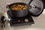 A black Ironlite Casserole filled with assorted vegetables and meat cooks on a digital induction cooktop at 268°F. The pot’s lid rests nearby, and utensils are placed to the left on the white countertop against a textured wall backdrop.