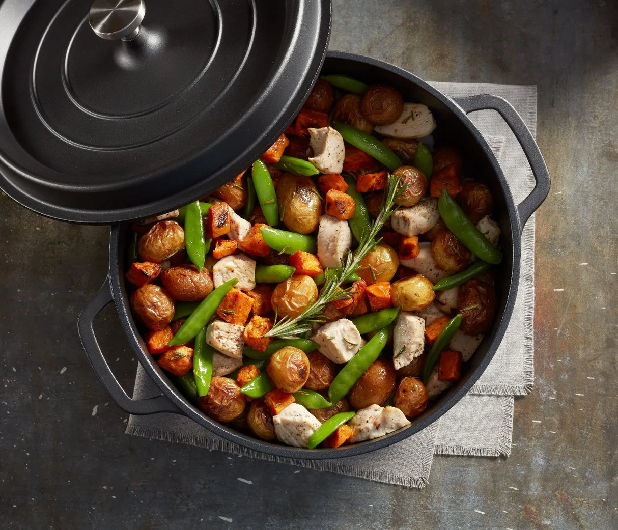 A black Ironlite Casserole filled with roasted baby potatoes, chicken pieces, snap peas, and diced carrots, garnished with rosemary. The non-stick casserole rests on a gray napkin atop a rustic table, its lid partially open.