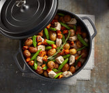A black Ironlite Casserole filled with roasted baby potatoes, chicken pieces, snap peas, and diced carrots, garnished with rosemary. The non-stick casserole rests on a gray napkin atop a rustic table, its lid partially open.
