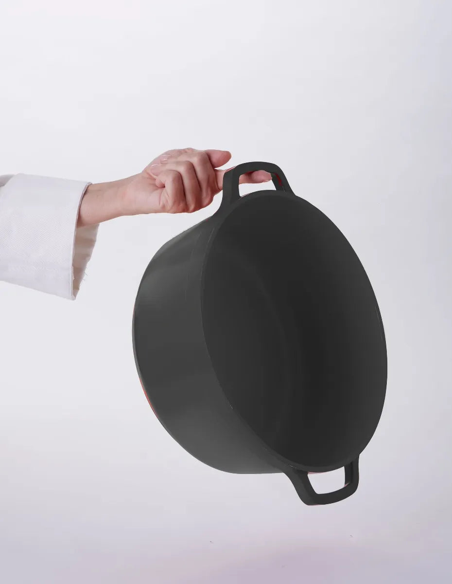 A person in a white sleeve holds a large Ironlite Casserole pot by the handle against a plain, light background. The cast iron pot, ideal for induction cooking, is angled toward the viewer with both handles visible.