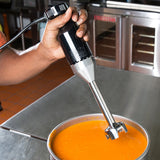 A person uses a light duty immersion blender to blend a pot of orange soup on a stainless steel counter in a commercial kitchen, with ovens and equipment visible in the background.