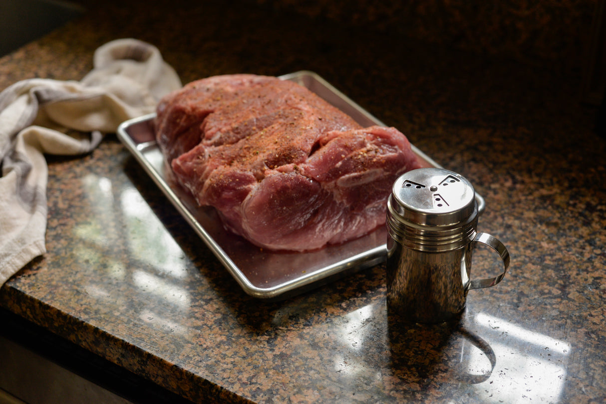 A large, raw pork roast seasoned with spices rests on a 1/4 size aluminized steel sheet pan atop a granite countertop. A metal shaker sits nearby, with a beige cloth loosely placed, all softly illuminated by sunlight from the left.