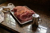 A large, raw pork roast seasoned with spices rests on a 1/4 size aluminized steel sheet pan atop a granite countertop. A metal shaker sits nearby, with a beige cloth loosely placed, all softly illuminated by sunlight from the left.