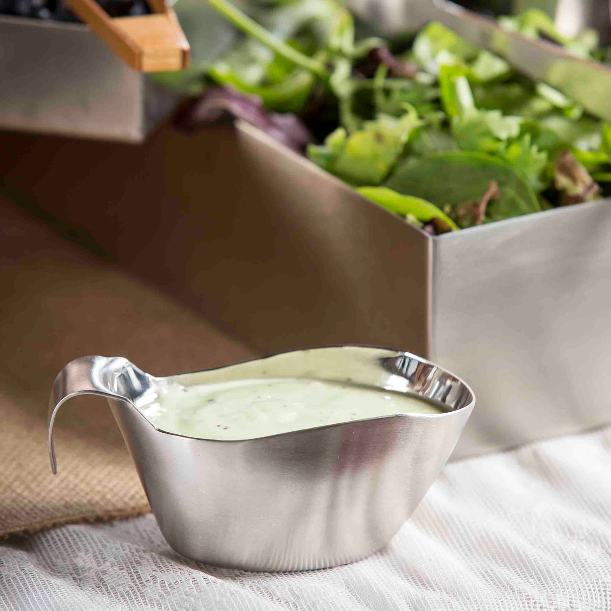 A stainless steel gravy boat filled with creamy green salad dressing sits on a white lace tablecloth. In the background, a metal container holds mixed leafy greens, slightly out of focus.
