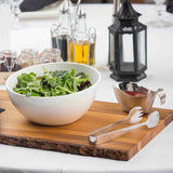 A white bowl of mixed leafy greens sits on a wooden board with stainless steel salad tongs, next to a small gravy boat with dressing. In the background are oil and vinegar bottles, jars, and a black lantern on a white tablecloth.
