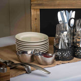 A stack of eight white plates sits on a burlap cloth next to stainless steel serving tongs, two 12 oz sauce dishes with red and dark sauces, and glass jars holding assorted forks, spoons, and knives in a black metal caddy.