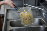 A hand holds a stainless steel fryer basket filled with raw, cut potato fries over a large sink. Water droplets drip from the fries and basket, indicating they have just been rinsed. The dishwasher safe basket sits near a faucet and metal counters in the background.