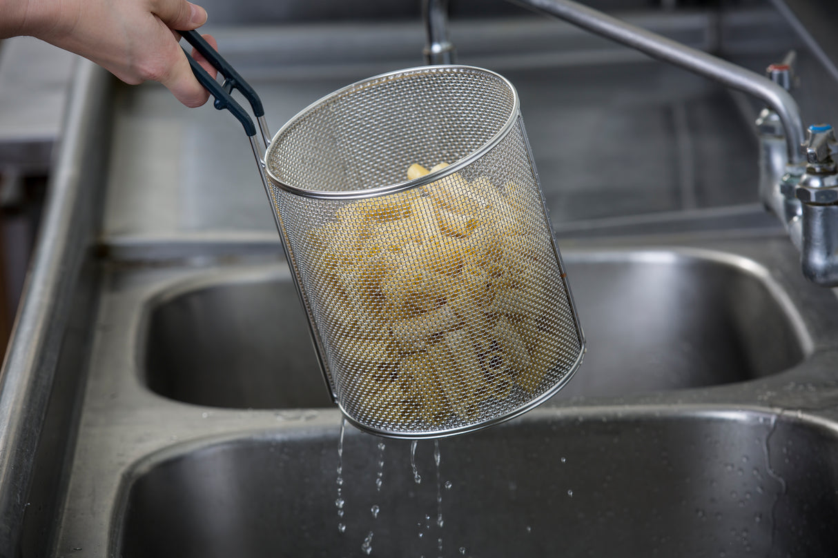 A hand holds a stainless steel pasta basket of cooked pasta above a sink, allowing water to drain. Water droplets drip from the dishwasher safe basket. In the background, a stainless steel double sink and faucet are visible.