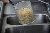 A hand holds a stainless steel pasta basket filled with cooked pasta above a kitchen sink, letting water drain out. The background features a sleek, multi-basin stainless steel sink and faucet. The pasta basket is dishwasher safe for easy cleaning.