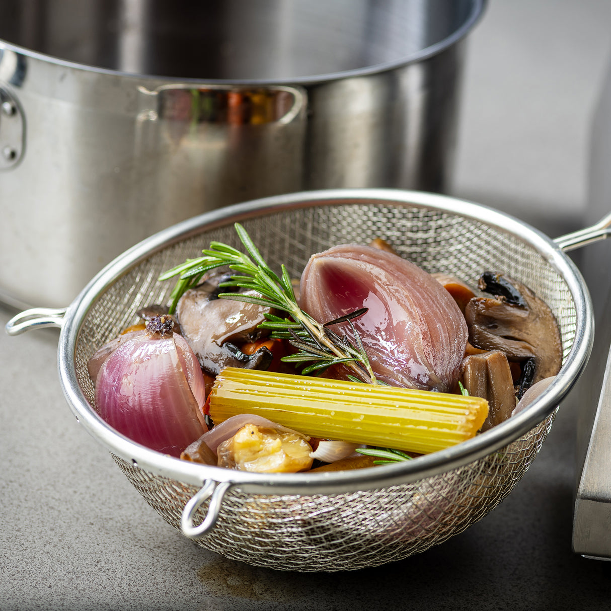 A metal strainer with a wooden handle and double medium mesh holds cooked vegetables—red onion, celery, mushrooms, and carrots—garnished with rosemary. The strainer rests on a countertop before a large stainless steel pot.