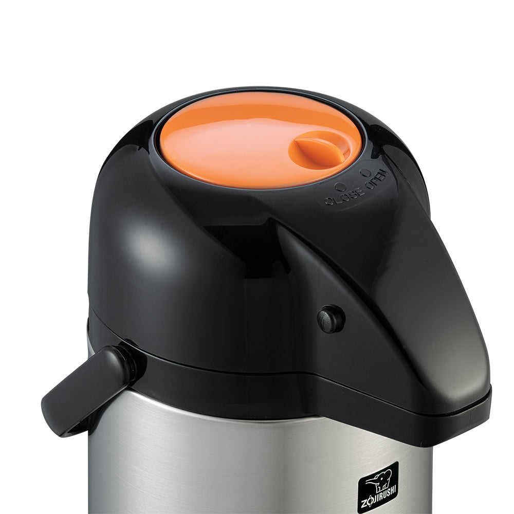 A close-up of a Zojirushi stainless steel airpot with a black lid and handle. The round lid features an orange top and spout, plus a black push-button for dispensing. Its stainless steel vacuum liner ensures excellent heat retention. The logo is visible on the front.