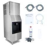 A stainless steel hotel ice machine stands on four legs, featuring a front ice dispenser and top cooling vents. Accessories include two coiled hoses, an instruction manual, a water filter, a pressure regulator with gauge, a white tube, and zip ties.