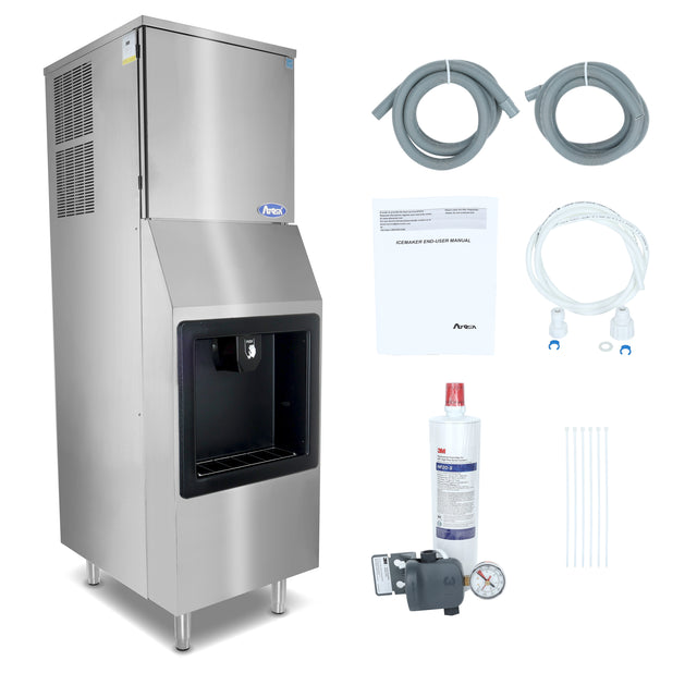 A stainless steel hotel ice machine stands on four legs, featuring a front ice dispenser and top cooling vents. Accessories include two coiled hoses, an instruction manual, a water filter, a pressure regulator with gauge, a white tube, and zip ties.