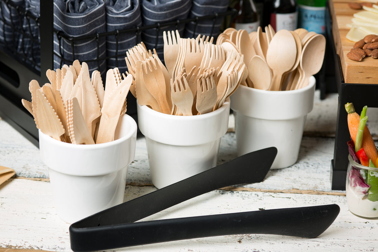 Three white ceramic cups hold wooden forks, knives, and spoons. The cups are side by side on a rustic white table. Heat safe stainless steel tongs lie in front of the cups, with napkins and various foods visible in the background.
