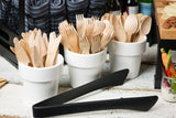 Three white ceramic cups hold wooden forks, knives, and spoons. The cups are side by side on a rustic white table. Heat safe stainless steel tongs lie in front of the cups, with napkins and various foods visible in the background.