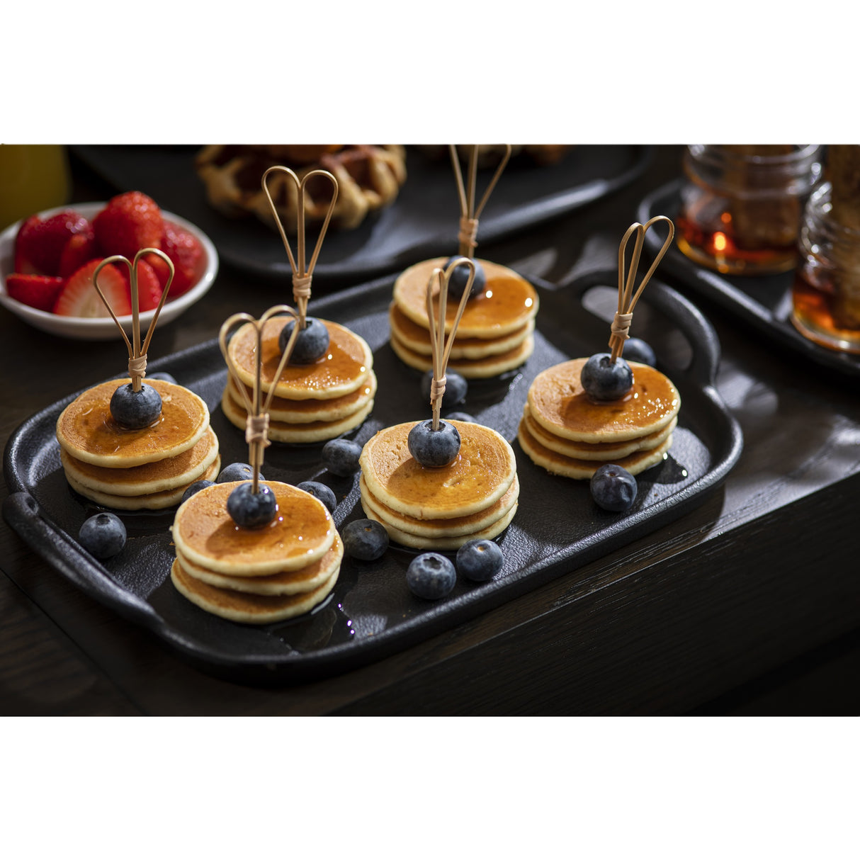 A black tray holds six stacks of mini pancakes topped with syrup, a Heart Looped Pick, and two blueberries each. Extra blueberries are scattered on the tray. In the background, TableCraft Products include cups, strawberries in a bowl, a waffle, and jars of syrup.