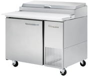 A commercial refrigerated stainless steel pizza prep table with a flat white work surface, hinged lid, single door with handle, ventilation grilles, and four caster wheels. The True brand is visible on the front door.