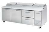 A large, three-section stainless steel Pizza Prep Table features a white cutting surface, three storage compartments, two pull-out drawers, overhead lid covers, and ventilation grates on the right, all mounted on caster wheels for easy mobility.