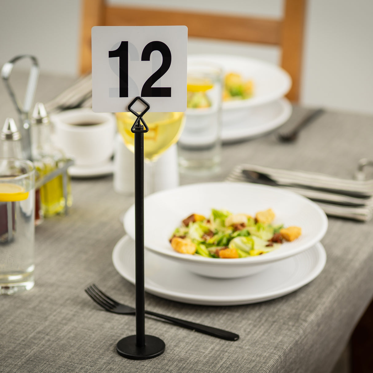 A restaurant table set for two with salads, drinks, utensils, and a TableCraft number holder displaying 12. The table features a gray tablecloth, wooden chair, and neatly arranged plates, glasses, and condiments for dining.