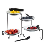 A three-tiered black powder coated metal stand holds four white bowls filled with cut pineapple, blueberries, strawberries, and mixed berries. Each bowl has a black serving tong or spoon. The stand is set on a white surface.