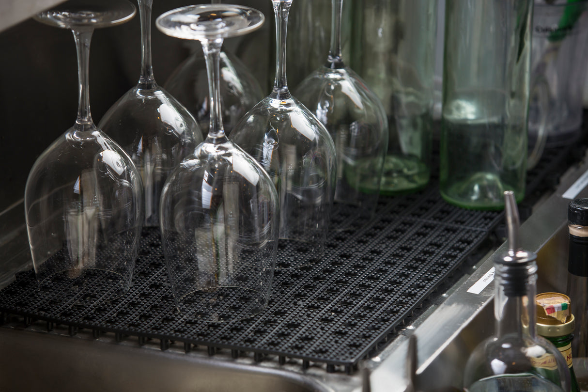 Five clear wine glasses are placed upside down on a black non-slip bar mat next to several tall green glass bottles. The scene is set in a kitchen or bar area, with various other bottles and containers visible around the edges.