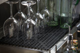 Five clear wine glasses are placed upside down on a black non-slip bar mat next to several tall green glass bottles. The scene is set in a kitchen or bar area, with various other bottles and containers visible around the edges.
