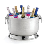 A large, round, 6-gallon stainless steel beverage tub with a hammered texture and side handles is filled with ice, chilling bottles of sparkling wine or champagne topped with colorful pink, blue, and gold foil.
