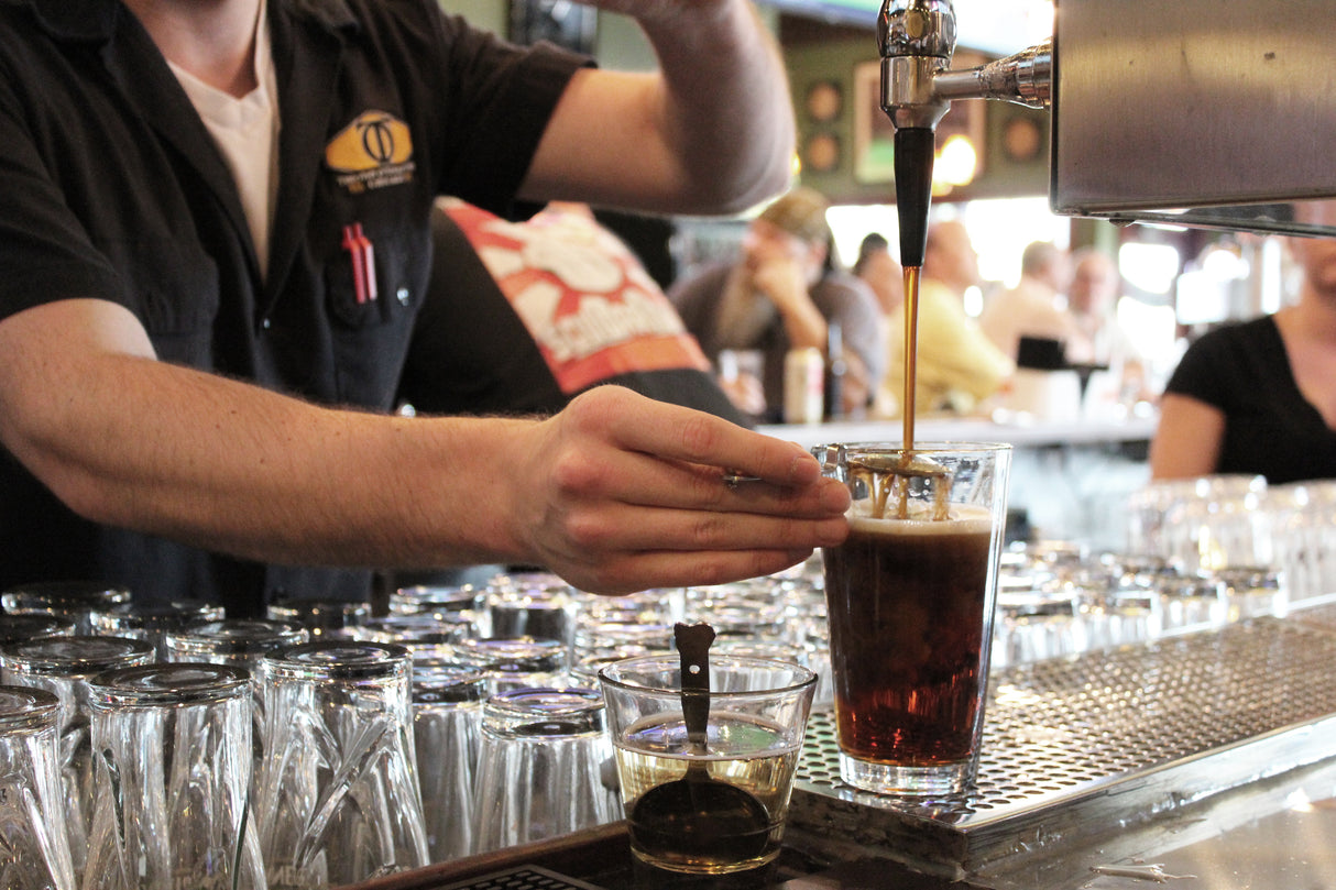 A bartender fills a pint glass with dark beer from a tap, carefully holding the glass at an angle and reaching for a stainless steel layering spoon. Several clean, dishwasher safe glasses are stacked nearby, with patrons visible in the blurred background.