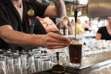 A bartender fills a pint glass with dark beer from a tap, carefully holding the glass at an angle and reaching for a stainless steel layering spoon. Several clean, dishwasher safe glasses are stacked nearby, with patrons visible in the blurred background.