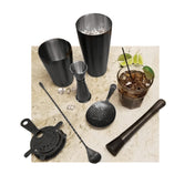 A marble countertop displays a Premium Barware Set: two stainless steel cocktail shakers, a jigger, muddler, strainer, long bar spoon, and a drink with ice, soda, lime wedges, and herbs. A few ice cubes are scattered on the surface.