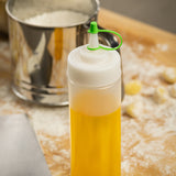 A clear plastic squeeze bottle with a tethered cap and standard cone tip tops, filled with yellow oil, sits on a floured wooden surface. In the background are a metal flour sifter and small pieces of dough, with flour scattered across the scene.
