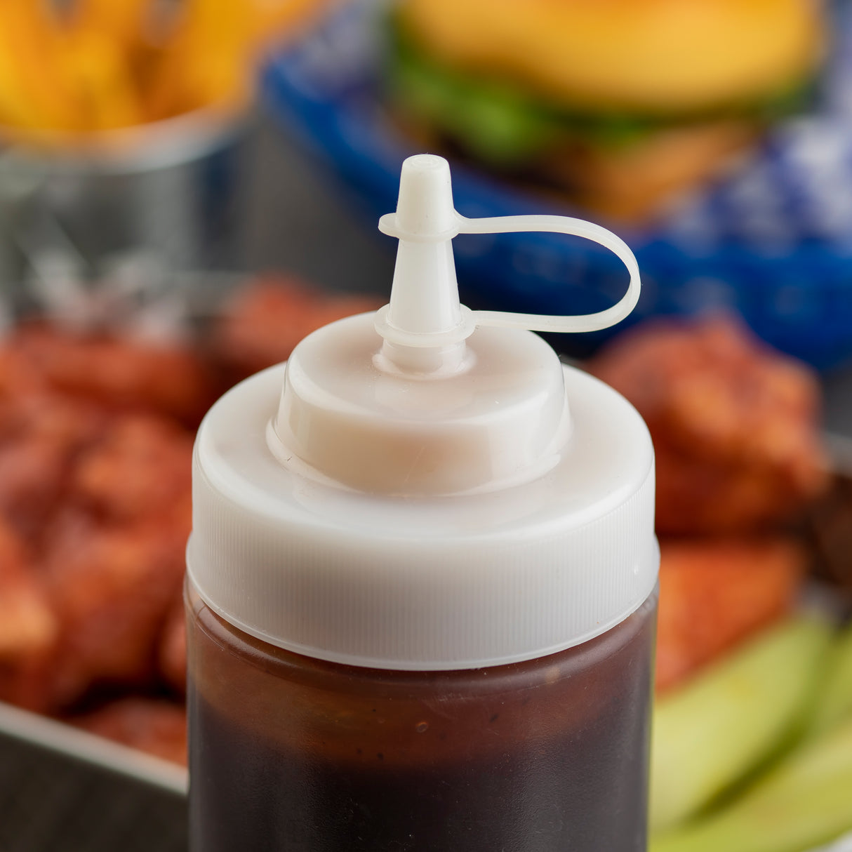 A close-up of a plastic squeeze bottle with a white, dishwasher safe cap and flip-top nozzle filled with dark brown sauce. In the blurred background are crinkle-cut fries, a hamburger in a blue basket, sauced wings, and sliced pickles.