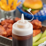 A close-up of a plastic squeeze bottle with a white, tethered cap and attached lid, filled with brown sauce. In the blurred background, French fries, a hamburger, fried chicken, and pickle spears sit on a metal tray.